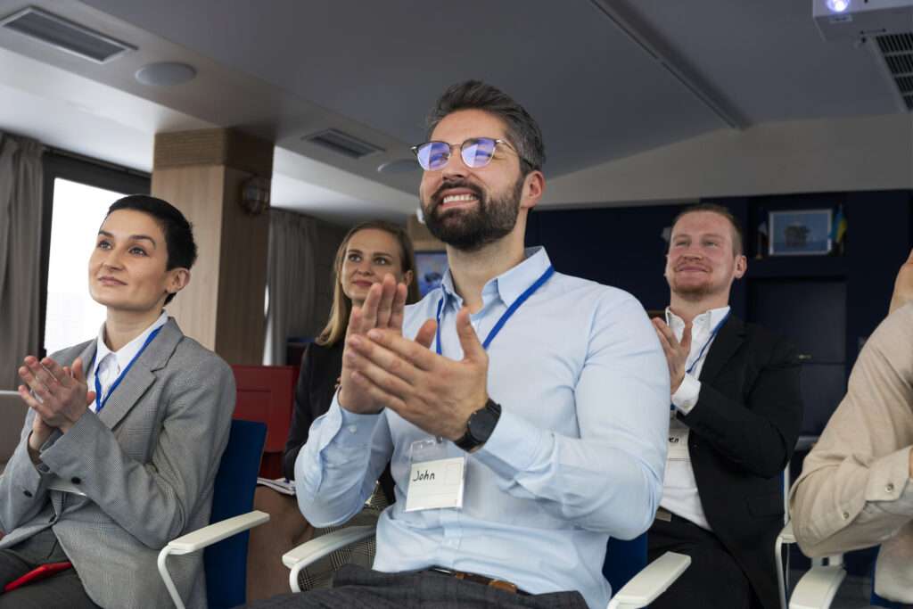 Grupo de personas con vestimenta formal aplaude a un ponente que se presentó en un evento corporativo. 